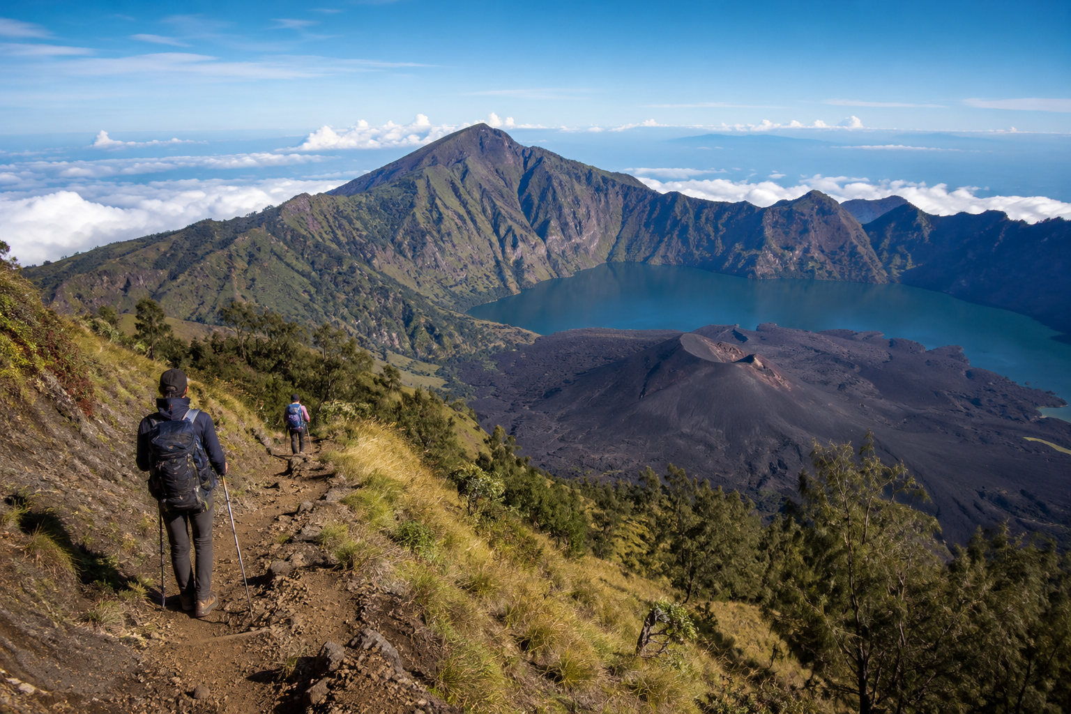 Hikers trekking the Mount Rinjani route with views of Segara Anak crater lake and volcanic terrain in Lombok, Indonesia.